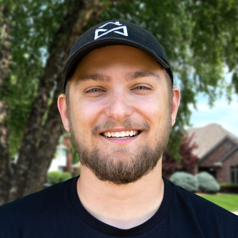 Smiling man wearing a cap in front of a house surrounded by greenery.