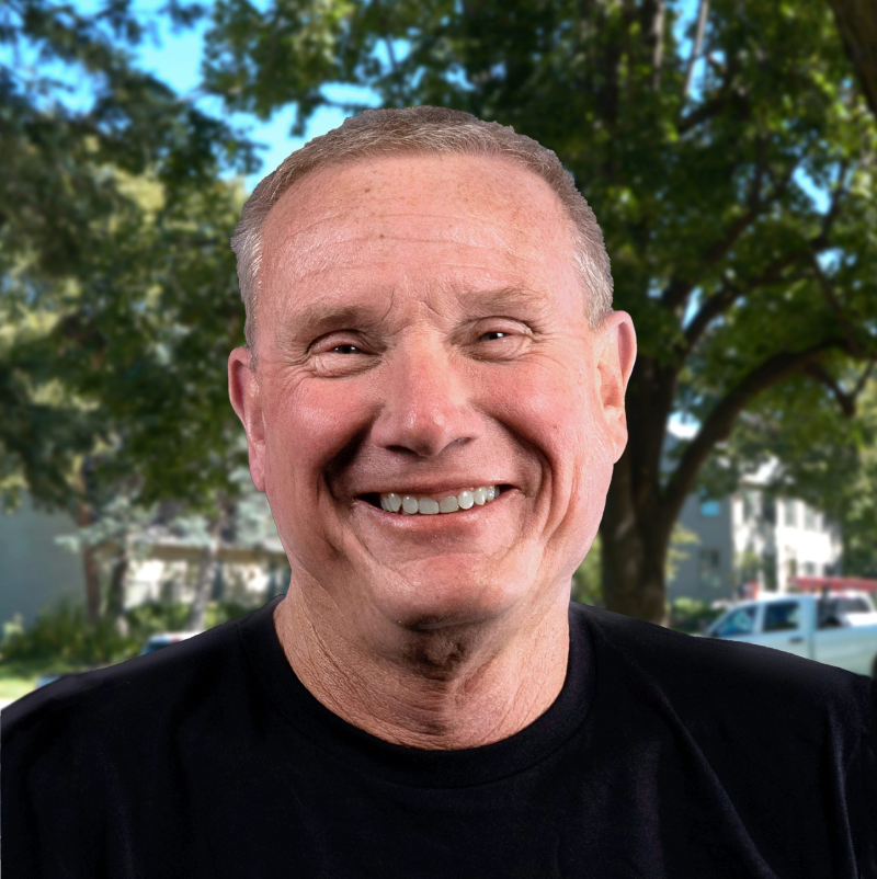 Smiling man stands outside with trees and buildings in the background.
