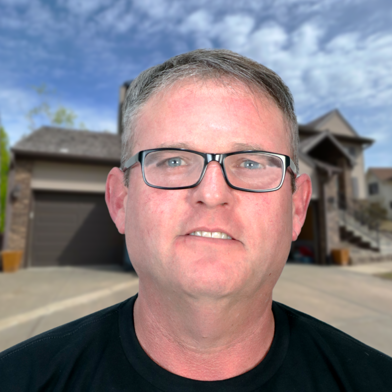 Man in glasses standing in front of a suburban house under a blue sky.