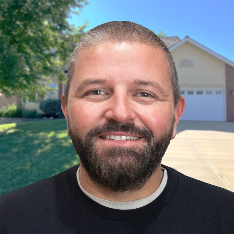 A smiling man stands in front of a suburban house on a sunny day.