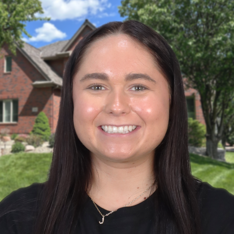 A smiling person stands in front of a brick house with a well-kept lawn.