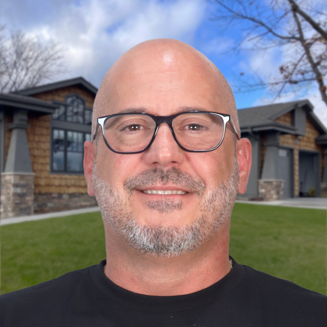 Man with glasses smiling in front of a modern house under a blue sky.