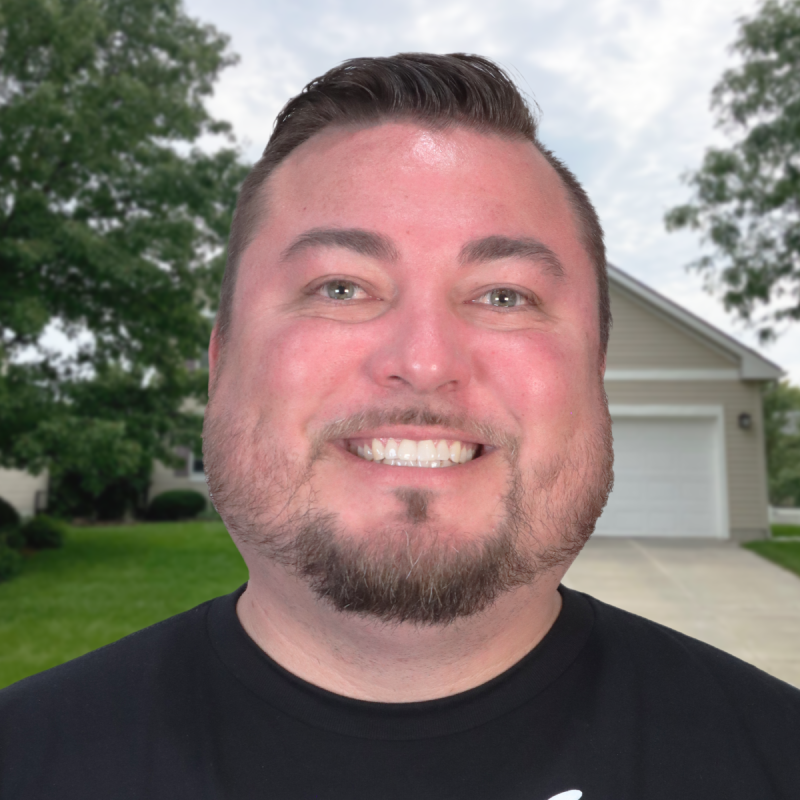Smiling man in front of a house and green lawn under a cloudy sky.